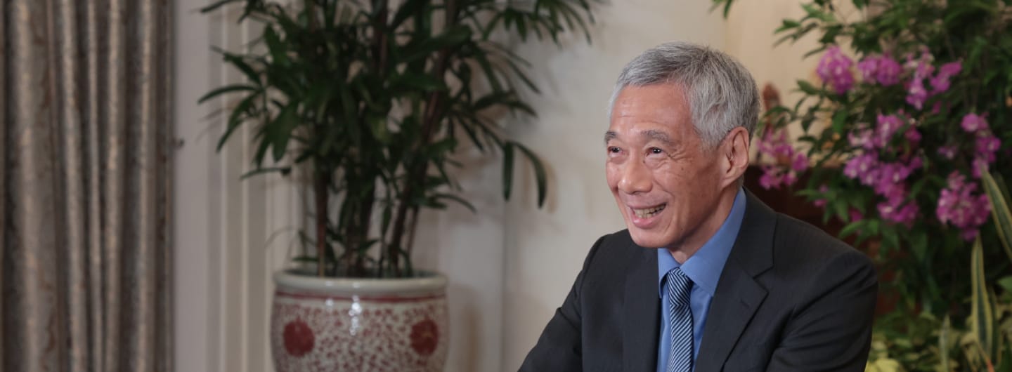 Lee Hsien Loong smiling, wearing a dark suit and striped tie, with plants and flowers.